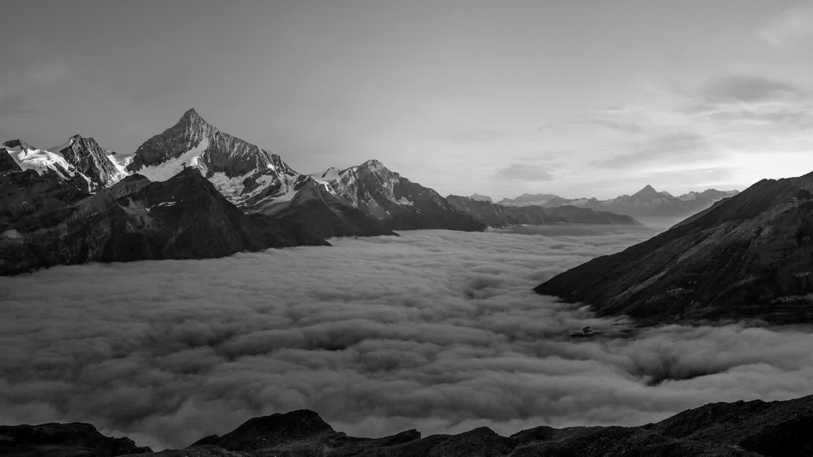 Black-and-white mountain peaks rising above a sea of valley fog, rocky ridge in the foreground