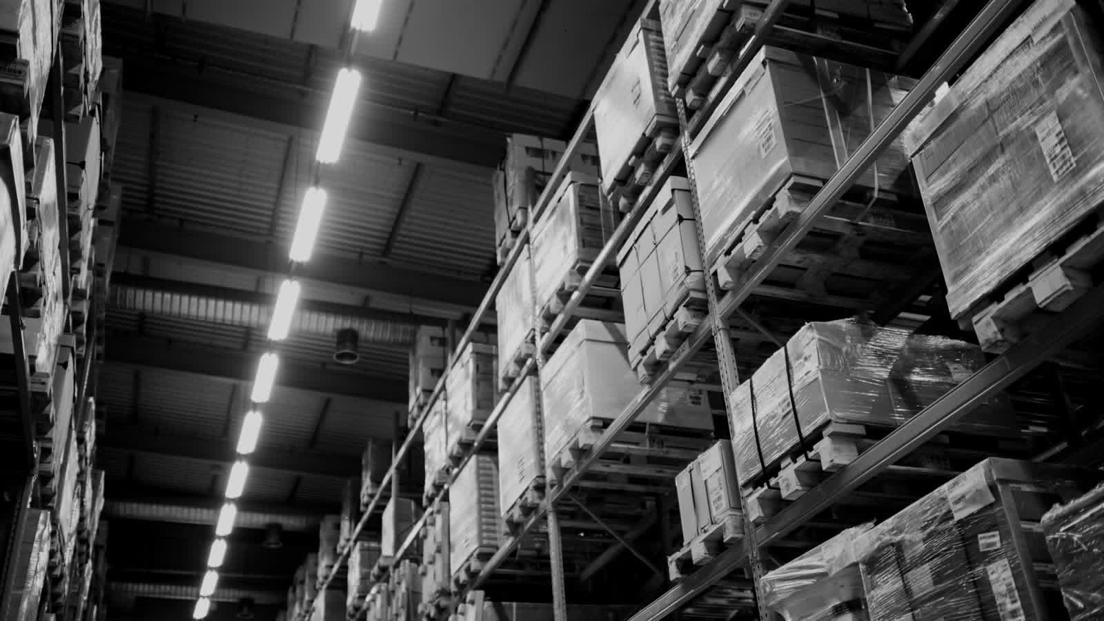 Black-and-white warehouse aisle with tall metal racks and shrink-wrapped pallets under overhead lights