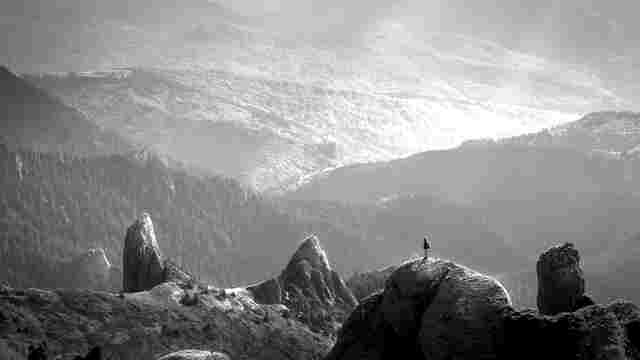 Black-and-white tiny figure on a jagged rock peak overlooking misty forested mountain ridges