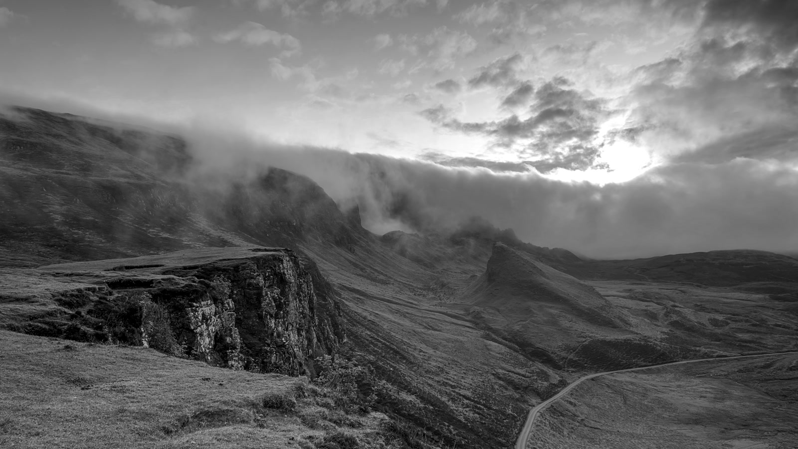 Black-and-white rocky cliff with fog cascading into a valley under a sunburst sky