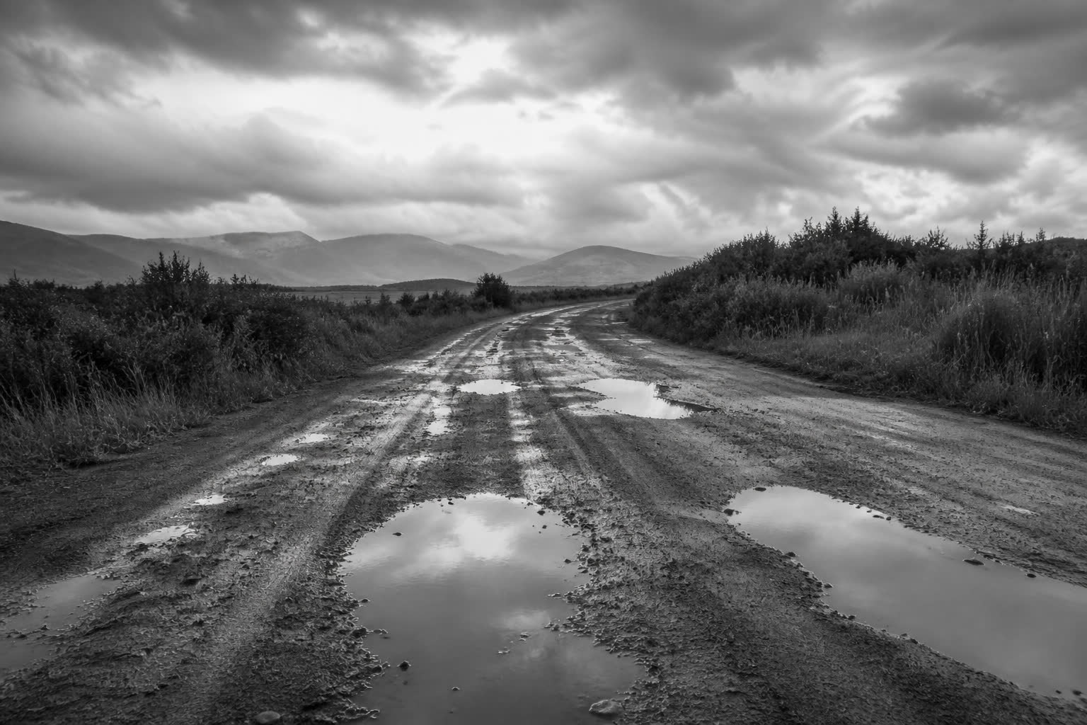 Black-and-white empty dirt road after rain, puddles reflecting sky