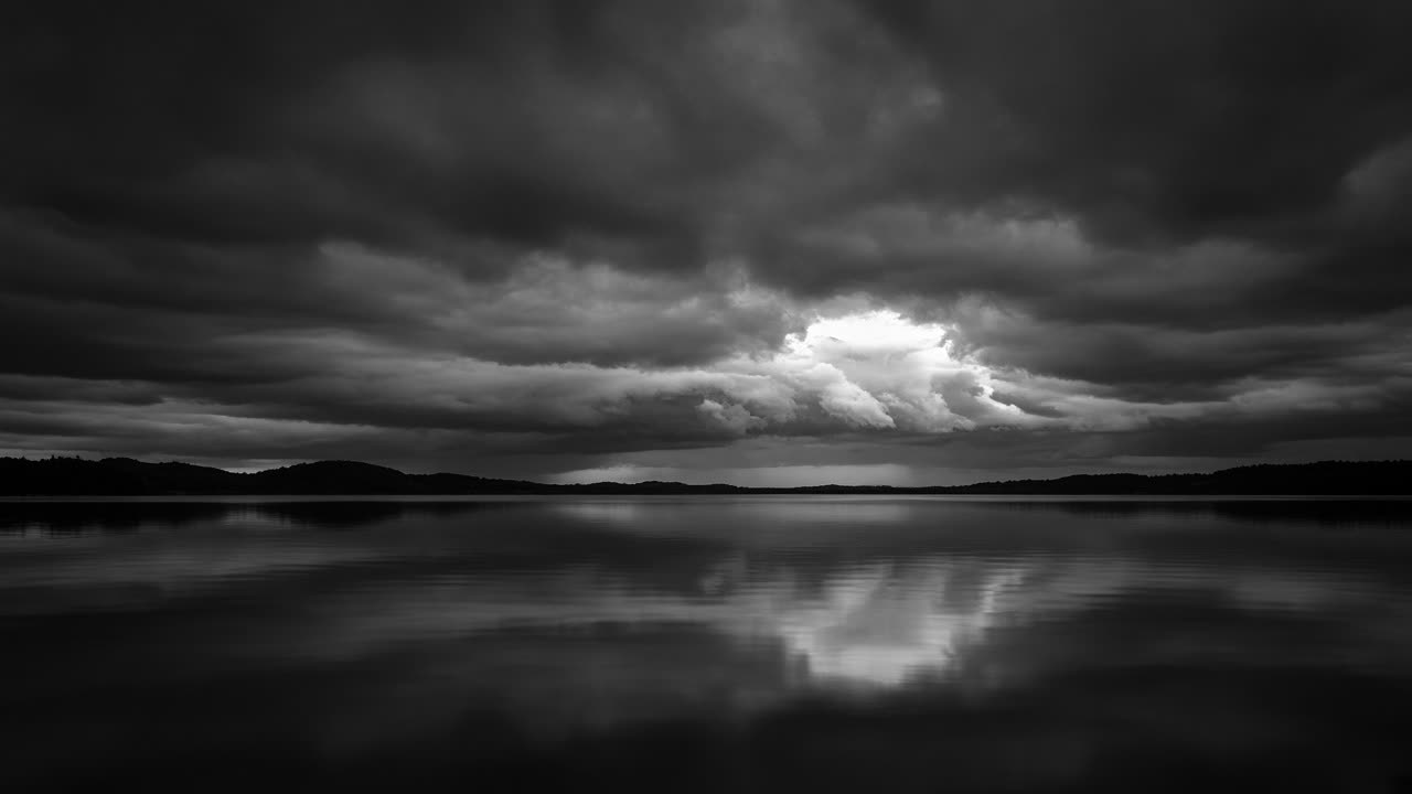 Black-and-white still lake under a storm sky with one break of light