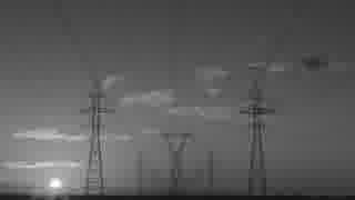 Black-and-white silhouette of high-voltage transmission towers and power lines toward a low sun