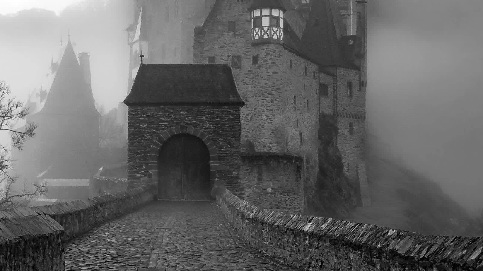 Black-and-white cobblestone bridge leading to a stone castle gatehouse in dense fog